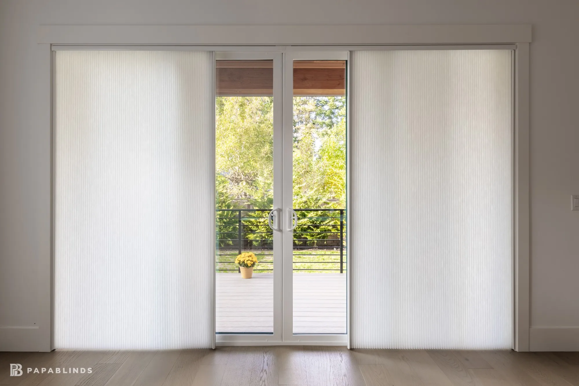 White vertical cellular shades on a sliding patio door, opened in the middle to the deck and backyard