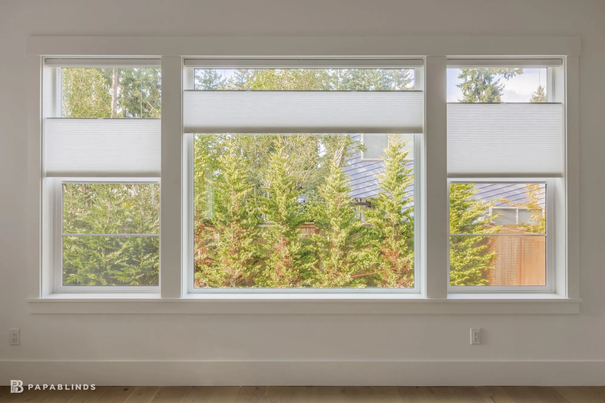 Three living room windows with white top-down bottom-up cellular shades lowered from the top to keep the view of the trees
